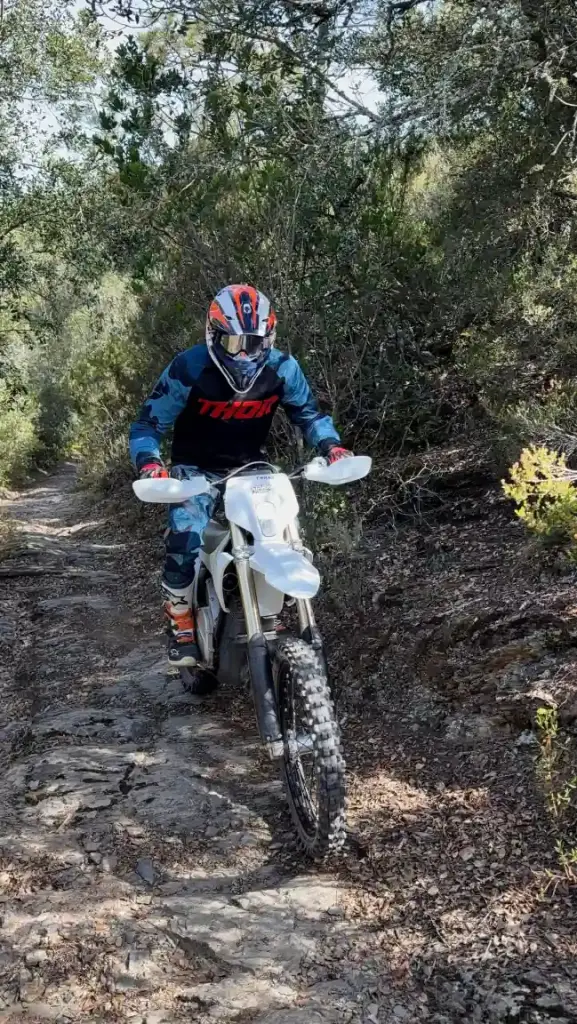 Un piloto baja con la moto eléctrica por un sendero rocoso entre la vegetación del bosque.